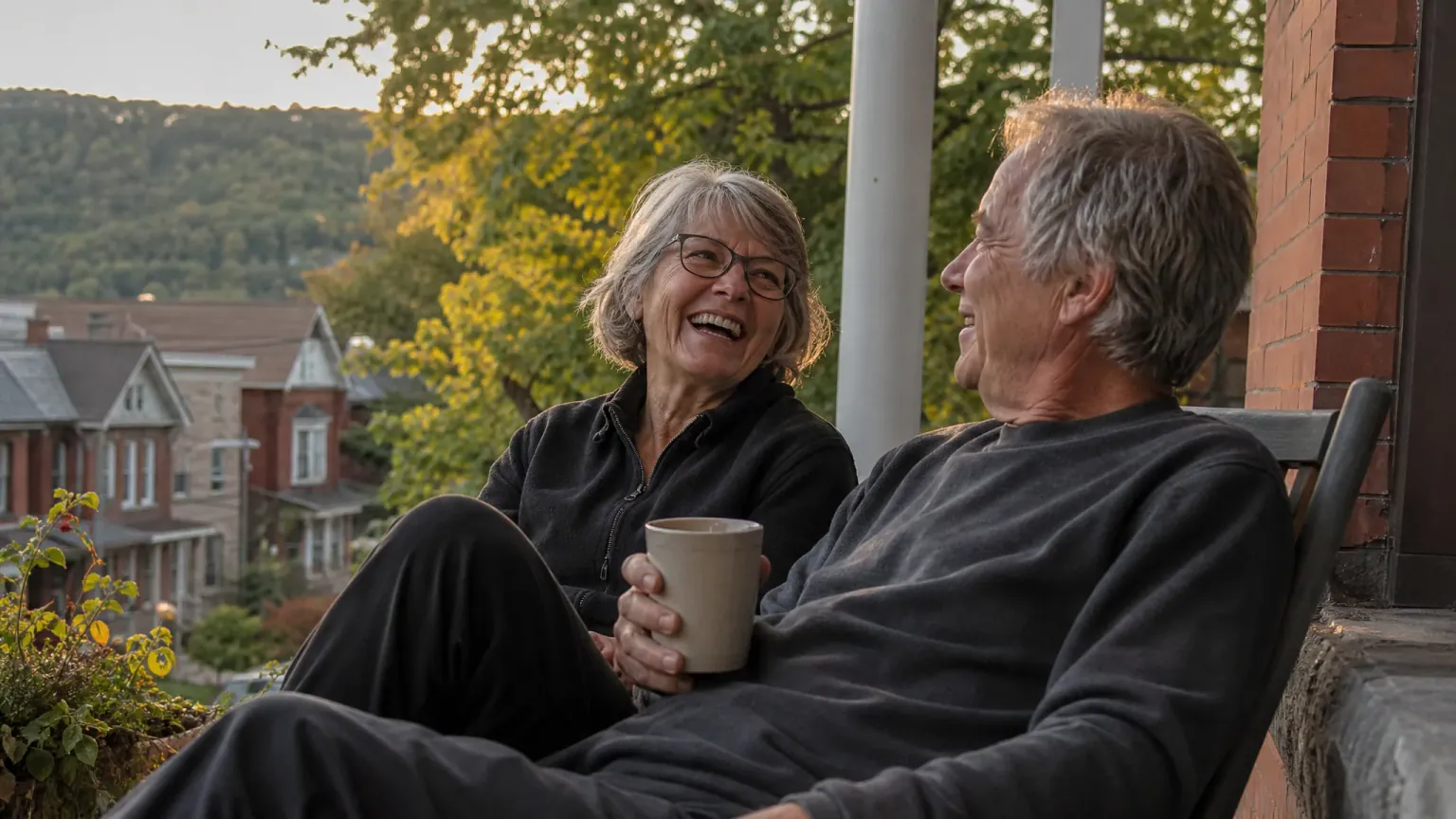 A Hamilton couple in their sixties laughing together on the front porch of their brick home at golden hour, the green escarpment rising behind the street