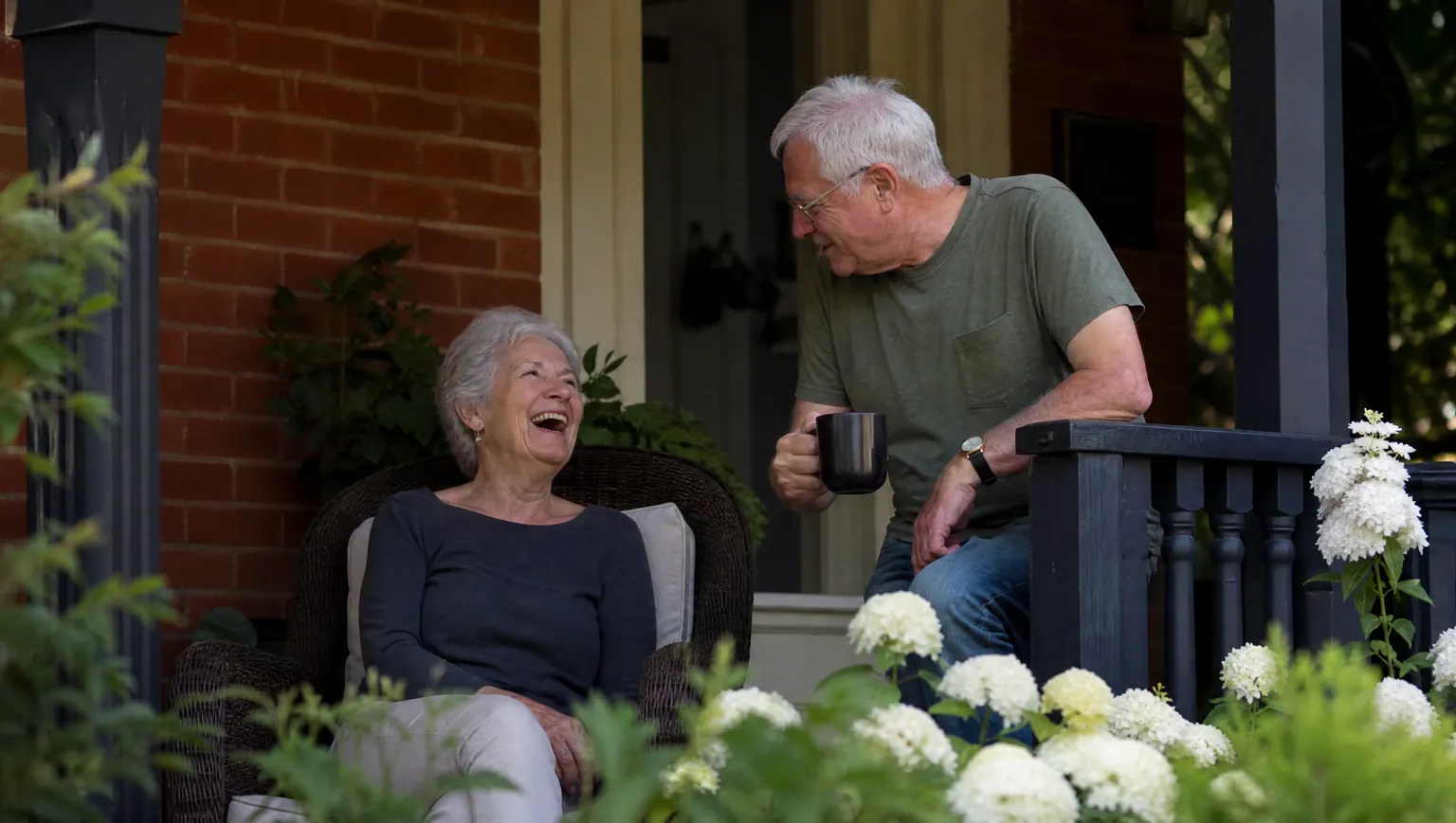 An older couple laughing together over coffee on the front porch of their brick home